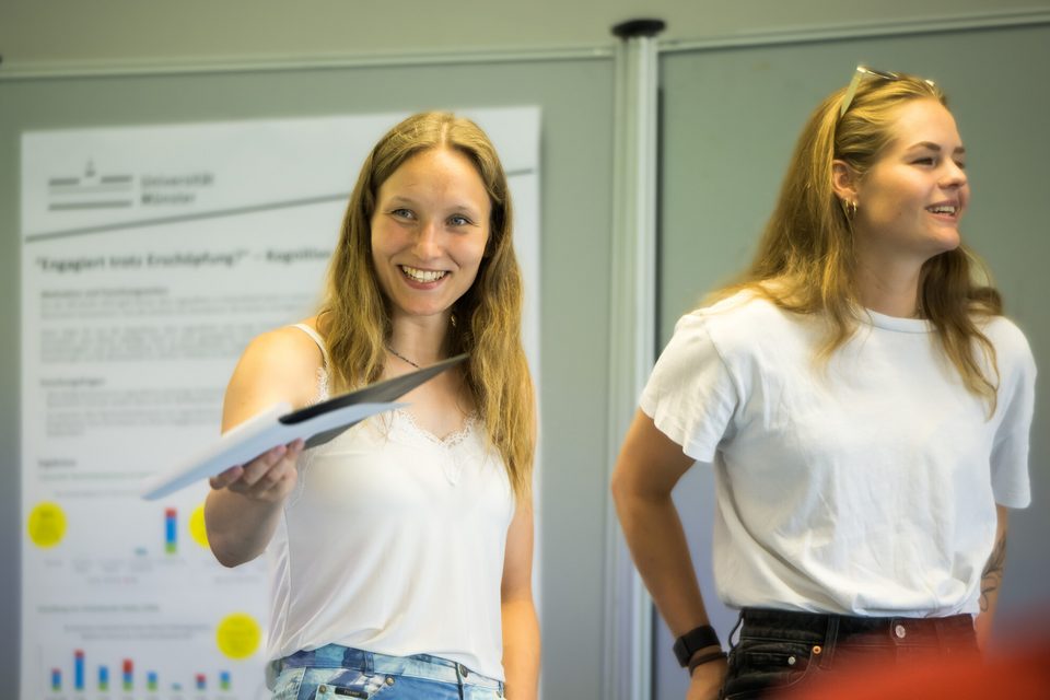 Zu sehen sind zwei junge Frauen. Sie tragen beide weiße Oberteile und haben lange blonde Haare. Die Frau auf der linken Seite hält eine Kladde in der rechten Hand und hält sie in die Luft. Die Frau auf der rechten Seite schaut nach rechts aus dem Bild. Sie hat eine Sonnenbrille in ihren Haaren. Ihre Hände hat sie in die hinteren Hosentaschen ihrer dunklen Jeans gesteckt.