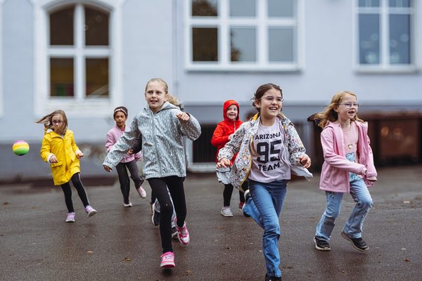 Sechs Kinder rennen über den Schulhof. Sie tragen bunte Regenjacken.