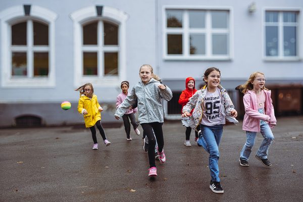 Sechs Kinder rennen über den Schulhof. Sie tragen bunte Regenjacken.