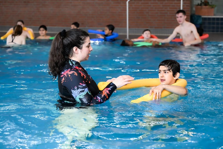 Zu sehen ist eine Situation im Schwimmbecken. Rechts ist ein Junge mit kurzen dunklen Haaren zu sehen, der eine gelbe Poolnudel um seinen Nacken gelegt hat. Vor ihm steht eine junge Frau mit einem langen Schwimmoberteil. Sie hat lange dunkle Haare, die sie in einen Pferdeschwanz zusammen gebunden hat. Sie erklärt dem Jungen etwas. Im Hintergrund sind weitere Kinder zu sehen.