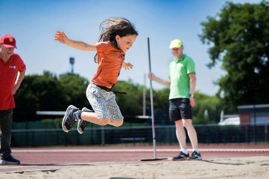 Zu sehen ist ein Mädchen mit langen dunklen Haaren. Sie ist gerade beim Weitsprung abgesprungen. Sie trägt ein orangefarbenes T-Shirt und eine kurze bunte Hose. Neben dem Weitsprungfeld stehen zwei Männer, die das Mädchen bei der richtigen Ausführung beobachten.